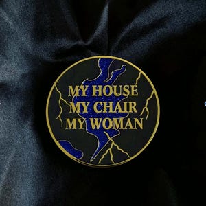 Peut inclure: Épinglette ronde noire et dorée avec l'inscription "MY HOUSE MY CHAIR MY WOMAN" en lettres dorées. L'épinglette présente un motif bleu pailleté et des éclairs dorés. L'épinglette est posée sur un fond en tissu noir.