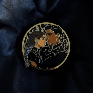 Op de afbeelding: Een ronde emaille pin met een gouden rand, met een paar in profiel. De figuren zijn zwart met bruine huidtinten en de vrouw heeft een blauwe vlecht. De pin heeft vlamachtige accenten en staat op een donkerblauwe achtergrond.