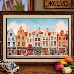 Op de afbeelding: Een ingelijste prent van kleurrijke Nederlandse huizen met winkels, een windmolenbeeldje, een houten schoen en een Holland-bord. De huizen zijn geschilderd in tinten blauw, oranje en wit. Een Nederlandse vlag is op de achtergrond.