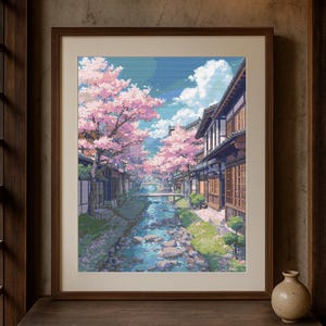 Puede incluir: Impresión artística enmarcada que representa una serena escena callejera japonesa. Los cerezos en flor rosas bordean un canal con un puente, edificios tradicionales y un cielo azul con nubes. La obra de arte se exhibe sobre una mesa de madera oscura.