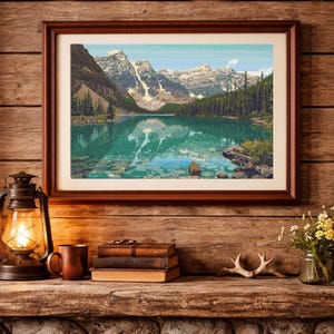 May include: A framed landscape print of a mountain lake scene with snow-capped peaks, displayed above a fireplace. The room features a rustic aesthetic with a lantern, books, and a mug on a wooden mantel. The fireplace has a stone facade.