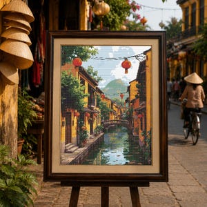Puede incluir: Un cuadro rectangular enmarcado sobre un caballete de madera que representa una escena de canal en una ciudad con edificios amarillos, farolillos rojos y un puente de piedra. La obra muestra árboles verdes, un canal y una montaña lejana.