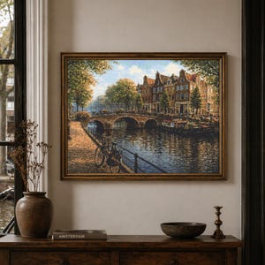 May include: A framed painting of an Amsterdam canal scene, showing a bridge, buildings, and boats. The artwork is in a gold frame and hangs on a neutral wall. A dark wooden cabinet with decor is in the foreground.