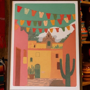May include: A framed print of a colorful street scene with buildings in shades of pink, yellow, and orange. A green cactus and string of festive flags add to the vibrant atmosphere. The artwork is displayed on a wooden easel.