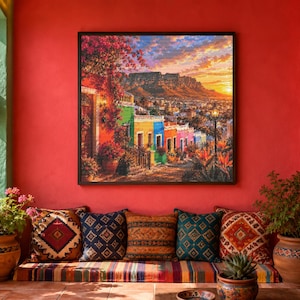 Peut inclure: Une impression d'art encadrée de forme carrée représentant une scène de rue animée avec des bâtiments colorés, des bougainvilliers en fleurs et une chaîne de montagnes sous un ciel au coucher du soleil, fixée sur un mur rouge vif au-dessus d'un banc.