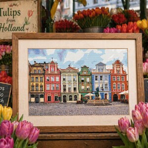 May include: A framed cross-stitch of a European city square, featuring buildings in yellow, red, green, and blue. The image is surrounded by bouquets of tulips, with signs reading "Tulips from Holland."