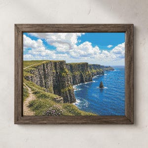 Puede incluir: Una impresión enmarcada de los Acantilados de Moher en Irlanda. La imagen muestra una vista panorámica de los acantilados, un sendero y el océano bajo un cielo azul con nubes blancas y esponjosas. El marco es de color marrón oscuro.