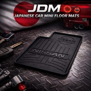 Op de afbeelding: Zwarte Nissan auto mini vloermat met de tekst "NISSAN" in een vetgedrukte lettertype. De mat heeft een gestructureerd oppervlak met parallelle lijnen. De afbeelding bevat ook de tekst "JDM JAPANESE CAR MINI FLOOR MATS".