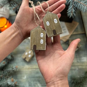 Puede incluir: Dos adornos de casa de madera con revestimiento marrón texturizado, detalles de ventanas blancas y puertas marrones se sostienen en las manos de una persona. Cada casa tiene un lazo de cuerda para colgar. El fondo incluye una superficie de madera y ramas de coníferas.
