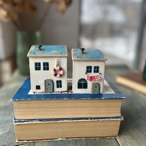 Może przedstawiać: Dwa miniaturowe białe domki z niebieskimi dachami, każdy z motywem marynistycznym. Jeden ma koło ratunkowe, drugi czerwoną i białą szachownicę. Stoją na stosie starych książek.