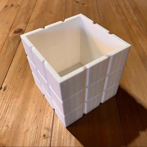 Può includere: Un vaso quadrato bianco con un design geometrico strutturato. Il vaso ha un interno cavo ed è appoggiato su una superficie di legno. Il vaso è progettato per uso interno o esterno.