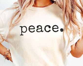 Puede incluir: Camiseta de color marfil con la palabra "peace" impresa en negro. La camiseta tiene mangas cortas y un corte relajado. El texto está en una fuente simple y en negrita. La camiseta se combina con vaqueros de mezclilla azul claro.