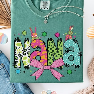 May include: A light green t-shirt featuring the word "NANA" in a colourful, patterned design. The design includes a pink bow, stars, and Easter-themed graphics. A silver necklace is draped across the shirt. The shirt is folded on a white surface with denim jeans and Easter decorations.