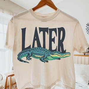 An ivory boxy crop top hanging on a wooden hanger, features a detailed green and teal alligator graphic under the bold black word LATER.