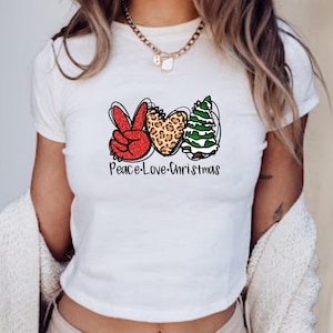 Puede incluir: Una camiseta blanca de manga corta con un diseño gráfico que incluye un signo de paz rojo con purpurina, un corazón con estampado de leopardo y un árbol de Navidad verde. Debajo, el texto Peace-Love-Christmas en letra negra.