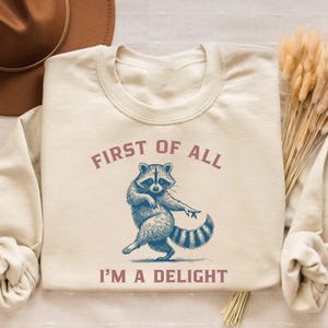 Sand-colored sweatshirt with a raccoon graphic. The raccoon is depicted in blue and white, with the text "FIRST OF ALL I'M A DELIGHT" in a mauve color. The sweatshirt is laid flat with a brown hat and dried flowers.