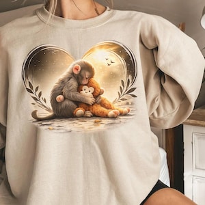 Sand-colored sweatshirt featuring a heart-shaped design with two monkeys hugging. One monkey is gray, the other is brown. The design includes a sunset background and the word "SAND" at the top.