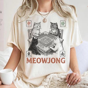 May include: Ivory-colored t-shirt featuring a black and white illustration of four cats playing Mahjong. The word "MEOWJONG" is printed in a brown, bold font below the table. The shirt has short sleeves and a relaxed fit.
