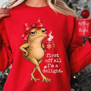 Puede incluir: Una sudadera de color rojo brillante con el gráfico de una rana verde texturizada que lleva un gran lazo rojo y sostiene una taza roja con un diseño de copo de nieve blanco. El texto dice: "first off all I'm a delight."
