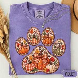 May include: A violet crewneck t-shirt featuring a large graphic of a paw print composed of five oval frames. Each frame contains a pumpkin with a plaid bow, autumn leaves, and hearts. The shirt has a Comfort Colors label at the inner neckline.