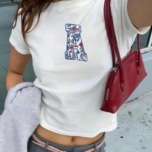 May include: White t-shirt with a floral embroidered "A" in blue and red. A red handbag with a zipper closure hangs from the shoulder. The person is wearing gray pants and a pink belt. A gray sweatshirt is held in the left arm.