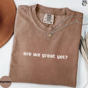 May include: Espresso-colored t-shirt with the phrase "are we great yet?" in white block letters. The shirt has a crew neck and a small tag at the neckline. A silver necklace with a round pendant is draped across the shirt.