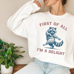 White sweatshirt with the text "FIRST OF ALL I'M A DELIGHT" and a dancing raccoon graphic. The raccoon is in shades of blue and gray.