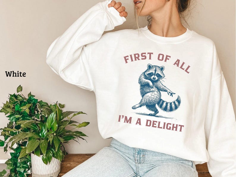 White sweatshirt with the text "FIRST OF ALL I'M A DELIGHT" and a dancing raccoon graphic. The raccoon is in shades of blue and gray.