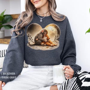 Heather dark gray sweatshirt featuring a heart-shaped graphic of a monkey hugging a teddy bear. The design includes a warm, golden background with leaves and birds.