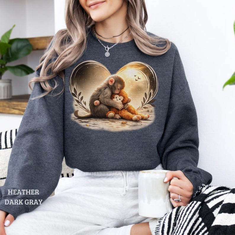 Heather dark gray sweatshirt featuring a heart-shaped graphic of a monkey hugging a teddy bear. The design includes a warm, golden background with leaves and birds.