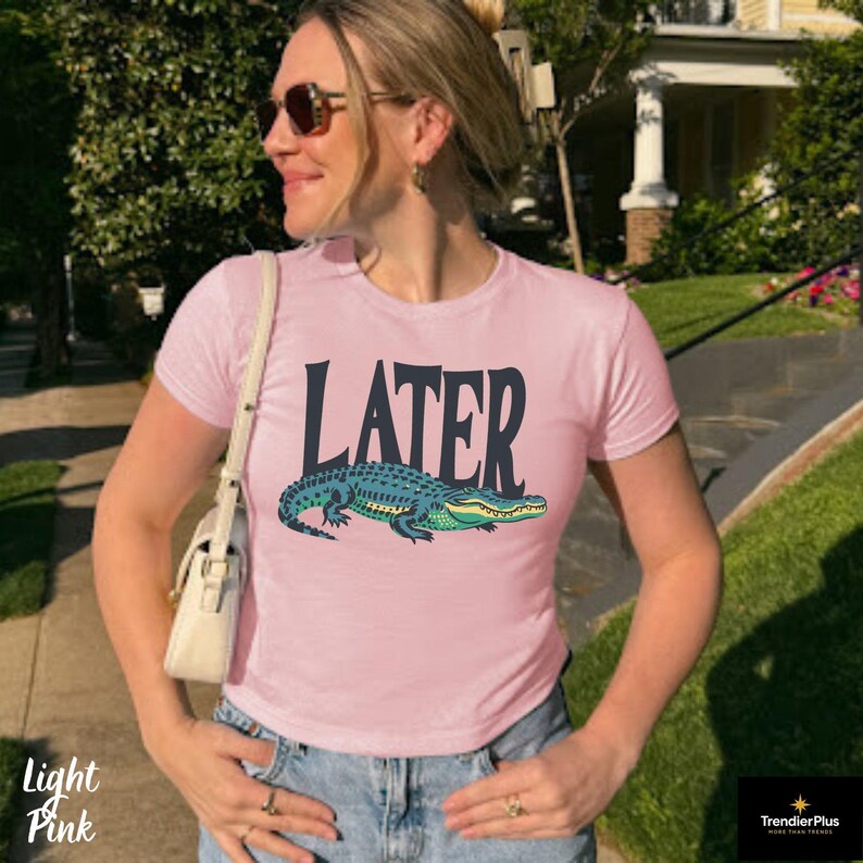 May include: A light pink short-sleeved t-shirt featuring the word LATER in large dark blue letters above a graphic illustration of a green and blue alligator. The shirt has a crew neckline and is worn with a white shoulder bag and blue jeans.