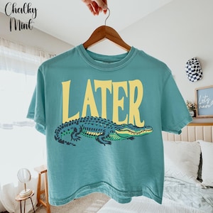May include: A teal short-sleeve t-shirt featuring the word LATER in large yellow capital letters above a graphic illustration of a green and dark blue alligator. The shirt is displayed on a wooden hanger against a light-colored room background.