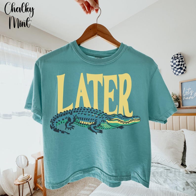 May include: A teal short-sleeve t-shirt featuring the word LATER in large yellow capital letters above a graphic illustration of a green and dark blue alligator. The shirt is displayed on a wooden hanger against a light-colored room background.