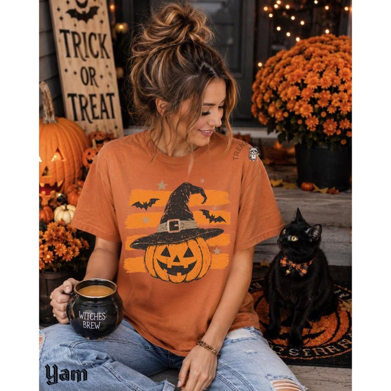Puede incluir: Una camiseta de manga corta color &oacute;xido con un gr&aacute;fico de una calabaza con sombrero de bruja, rodeada de murci&eacute;lagos y estrellas. La camiseta se combina con vaqueros azules y una taza negra con forma de caldero que dice Witches Brew.