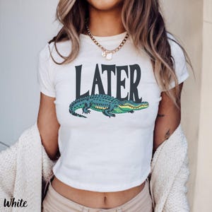 May include: A white short-sleeved t-shirt featuring the word LATER in large dark gray capital letters above a graphic illustration of a green and dark blue alligator. The design is centered on the chest of the garment.
