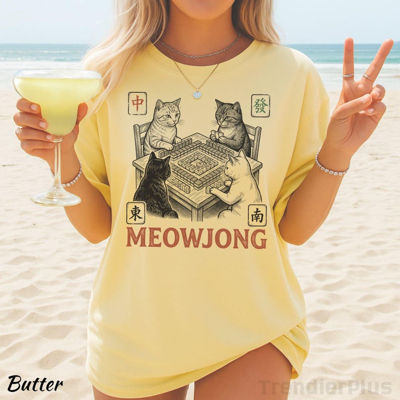 Może przedstawiać: Jasnoż&oacute;łty t-shirt z grafiką przedstawiającą cztery koty grające w mahjonga przy stole. Wz&oacute;r zawiera napis MEOWJONG oraz chińskie znaki w kwadratowych ramkach. Ilustracja ukazuje koty w r&oacute;żnych pozach podczas rozgrywki przy planszy.