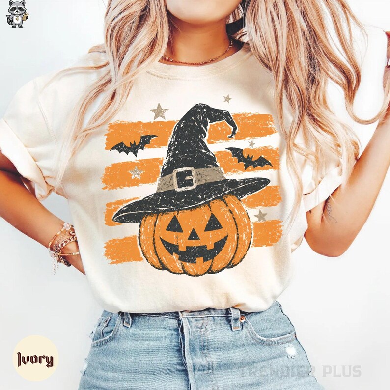 A model wearing an ivory t-shirt featuring a vintage jack-o'-lantern wearing a witch hat in front of distressed orange stripes and bats.