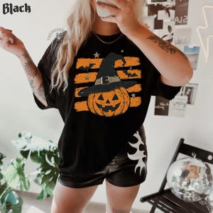 Puede incluir: Una camiseta negra con un estampado gr&aacute;fico de una calabaza naranja de Halloween con un sombrero de bruja negro. El dise&ntilde;o incluye pinceladas naranjas horizontales, estrellas peque&ntilde;as y siluetas de murci&eacute;lagos volando. La palabra Black aparece arriba.