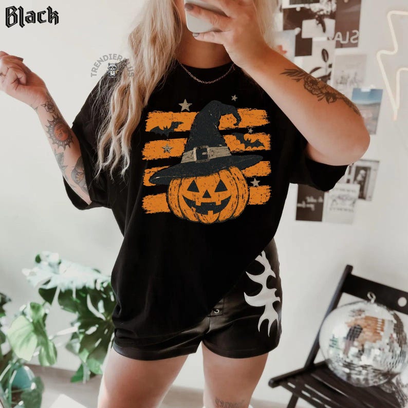 Puede incluir: Una camiseta negra con un estampado gr&aacute;fico de una calabaza naranja de Halloween con un sombrero de bruja negro. El dise&ntilde;o incluye pinceladas naranjas horizontales, estrellas peque&ntilde;as y siluetas de murci&eacute;lagos volando. La palabra Black aparece arriba.
