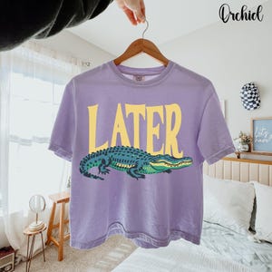May include: A light purple short-sleeve t-shirt featuring the word LATER in large yellow capital letters above a graphic illustration of a green and dark blue alligator. The shirt is displayed on a wooden hanger against a bright room setting.