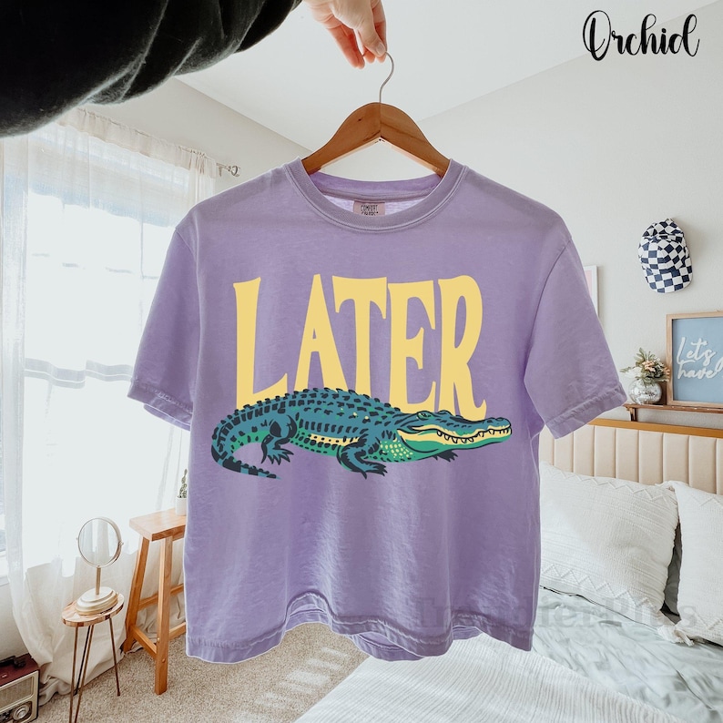 May include: A light purple short-sleeve t-shirt featuring the word LATER in large yellow capital letters above a graphic illustration of a green and dark blue alligator. The shirt is displayed on a wooden hanger against a bright room setting.
