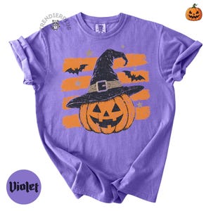 Puede incluir: Una camiseta violeta de manga corta con un gr&aacute;fico de una calabaza naranja que lleva un sombrero de bruja negro. El dise&ntilde;o incluye trazos de pincel naranjas horizontales, murci&eacute;lagos negros y estrellas peque&ntilde;as. El texto "TRENDIERPLUS" aparece cerca del cuello.