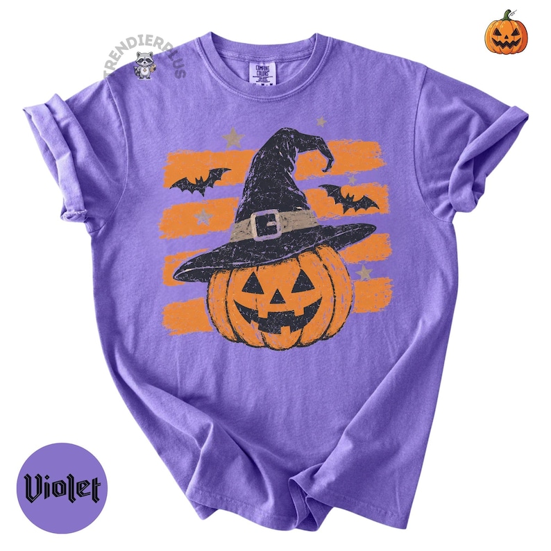 Puede incluir: Una camiseta violeta de manga corta con un gr&aacute;fico de una calabaza naranja que lleva un sombrero de bruja negro. El dise&ntilde;o incluye trazos de pincel naranjas horizontales, murci&eacute;lagos negros y estrellas peque&ntilde;as. El texto "TRENDIERPLUS" aparece cerca del cuello.