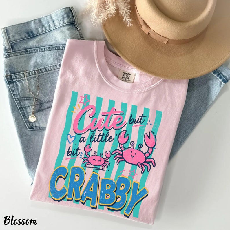 May include: A light pink t-shirt featuring a graphic print with teal vertical stripes, two pink crabs, and the text "Cute but a little bit CRABBY" in pink and blue lettering. The shirt is paired with light blue denim jeans and a tan hat.