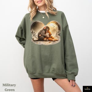 Military green sweatshirt featuring a heart-shaped graphic with a monkey hugging a teddy bear. The illustration has a warm, golden glow. The sweatshirt is a relaxed fit.