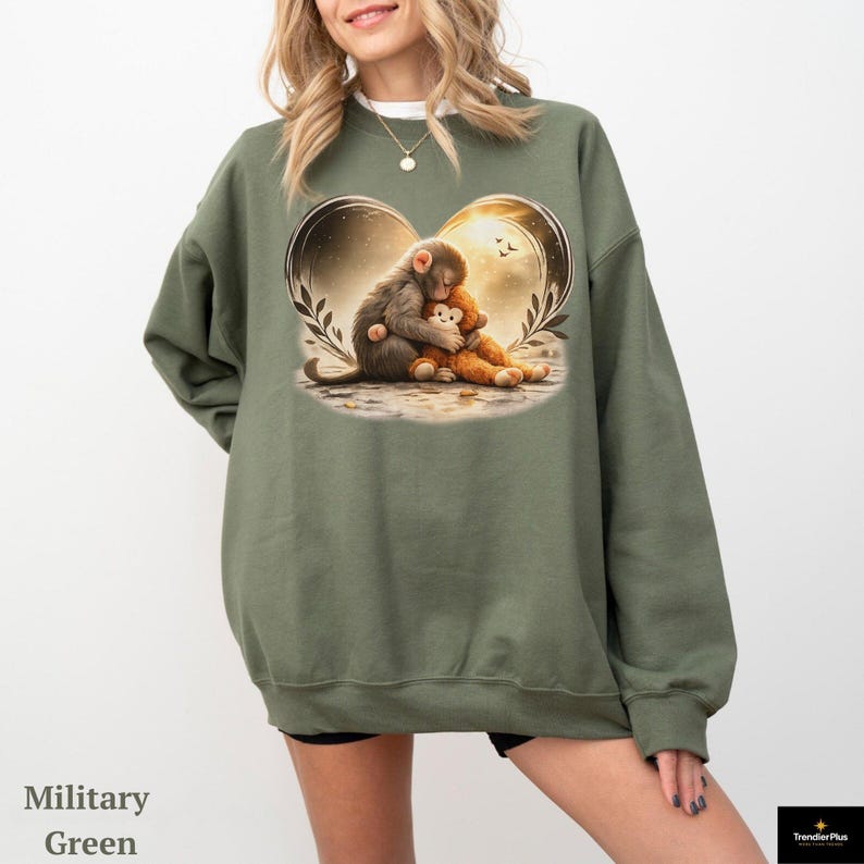Military green sweatshirt featuring a heart-shaped graphic with a monkey hugging a teddy bear. The illustration has a warm, golden glow. The sweatshirt is a relaxed fit.