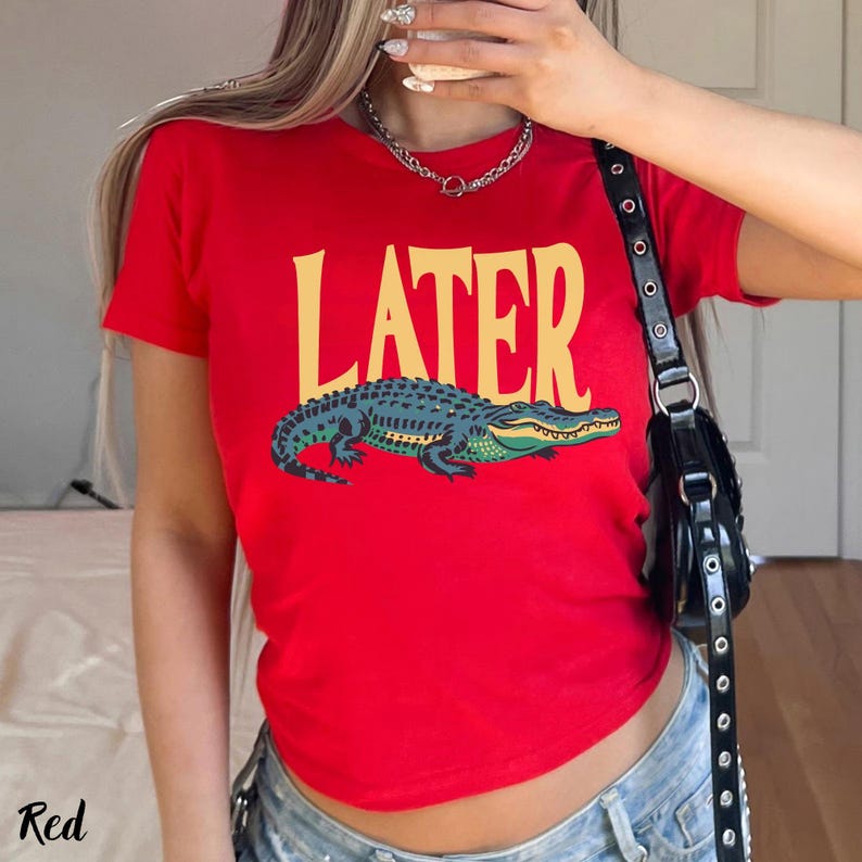 May include: A bright red short-sleeved t-shirt featuring the word LATER printed in large yellow capital letters above a graphic illustration of a green and dark blue alligator. The shirt has a crew neckline and a standard fit design.