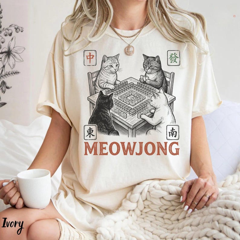 May include: Ivory-colored t-shirt featuring a black and white illustration of four cats playing Mahjong. The word "MEOWJONG" is printed in a brown, bold font below the table. The shirt has short sleeves and a relaxed fit.