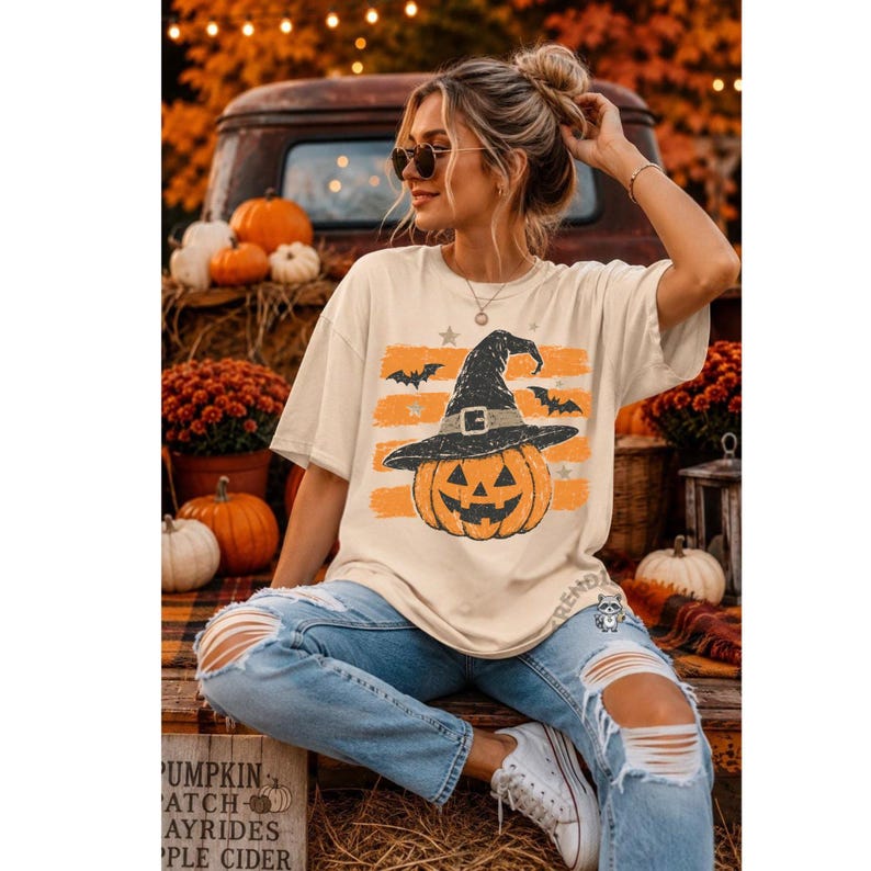 Puede incluir: Una camiseta de manga corta color crema con un gr&aacute;fico de una calabaza de Halloween con un sombrero de bruja negro, rodeada de rayas horizontales naranjas y peque&ntilde;as estrellas. La camiseta se combina con unos vaqueros azul claro desgastados.
