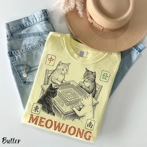 Może przedstawiać: Bladoż&oacute;łty t-shirt z grafiką przedstawiającą cztery koty grające w mahjonga przy stole. Wz&oacute;r zawiera chińskie znaki w kwadratowych ramkach oraz napis MEOWJONG dużymi czerwonymi literami, ułożony na jasnoniebieskich dżinsach.